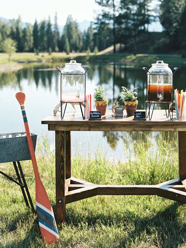 outdoor wedding drink station idea with glass dispensers of iced tea and lemonade on a wooden table in front of a lake