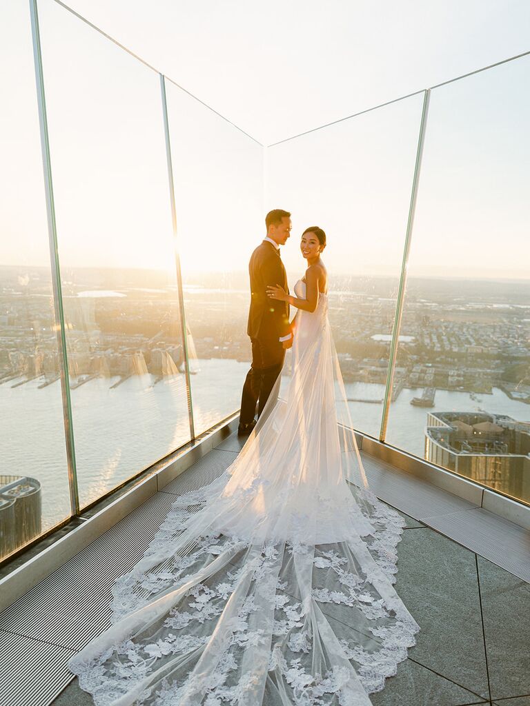 A happy couple stands on a glass promontory overlooking the big city, as the sun rises.