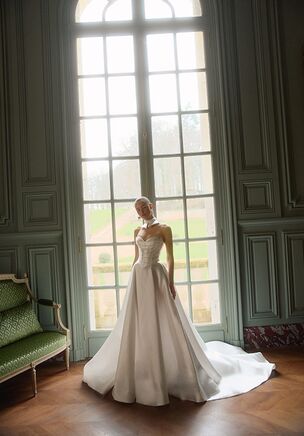 Luminous texture meets architectural geometry in this light ivory bridal creation from WONÃ Concept. Woven from structured Mikado and intricate beading, the gown frames the collarbone with a classic dipped neckline, highlighting the exposed stitching on the bodice. Scattered pearls lead to a dramatic opening in the skirt. An open back balances the sweep of a chapel-length train. A coordinating choker accessory transforms the neckline, offering a multi-piece presentation entirely ready to change.