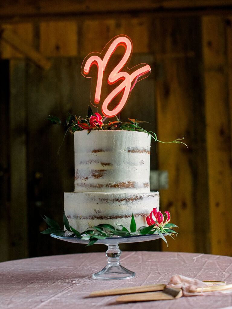 Rustic two-tier wedding cake with greenery and neon initial cake topper