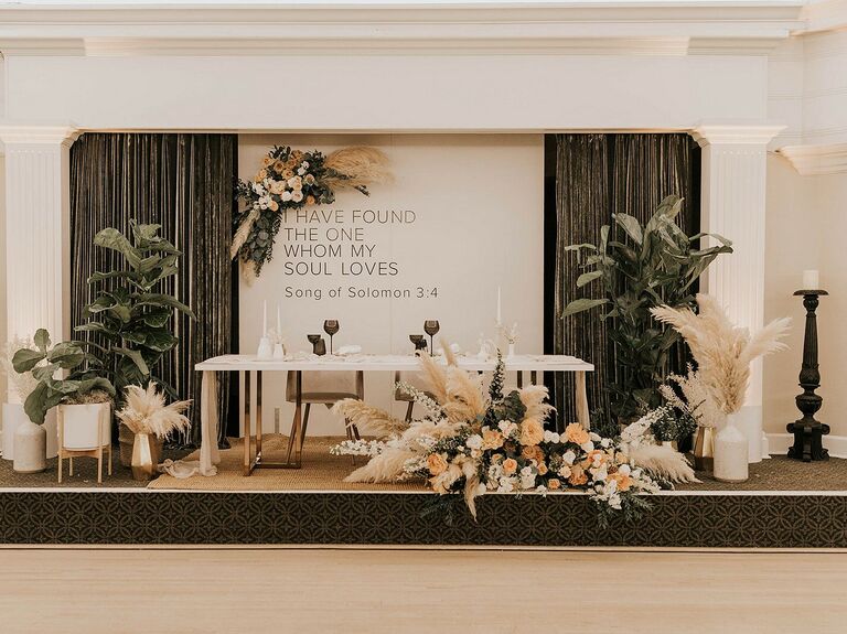indoor wedding sweetheart table idea with table on a raised platform decorated with pampas grass arrangements, potted plants and a backdrop with bible verse from song of solomons 3.4