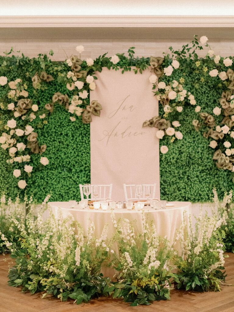 romantic wedding sweetheart table decor with greenery wall, white flowers and custom backdrop with couple's names written in calligraphy