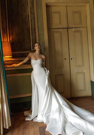 Artemis gown is a sophisticated mermaid silhouette in a soft light ivory shade. Crafted from delicate pearl-embellished lace, this strapless dress offers a refined and modern bridal look. The detachable mikado train adds a dramatic and regal touch, while the base dress remains minimalist and elegant without it — ideal for transitioning from ceremony to reception. The train is attached with discreet lacing, completely hidden beneath the fabric for a seamless appearance. A statement bow at the back waistline draws attention to the silhouette and adds a hint of romantic flair. The train extends into a graceful chapel length, completing the look with timeless sophistication.