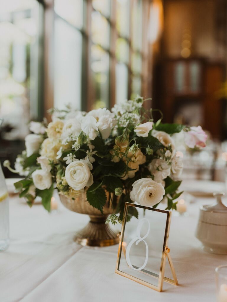 classic small wedding centerpiece with white roses, ranunculus and greenery in small gold compote vase with framed glass table number