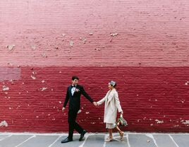 bride and groom hold hands walking outside against a pink and red brick painted wall
