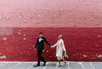 bride and groom hold hands walking outside against a pink and red brick painted wall