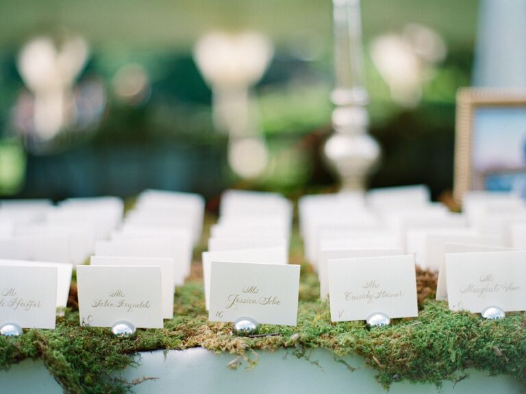 white escort cards with names written in gold calligraphy displayed atop a bed of moss