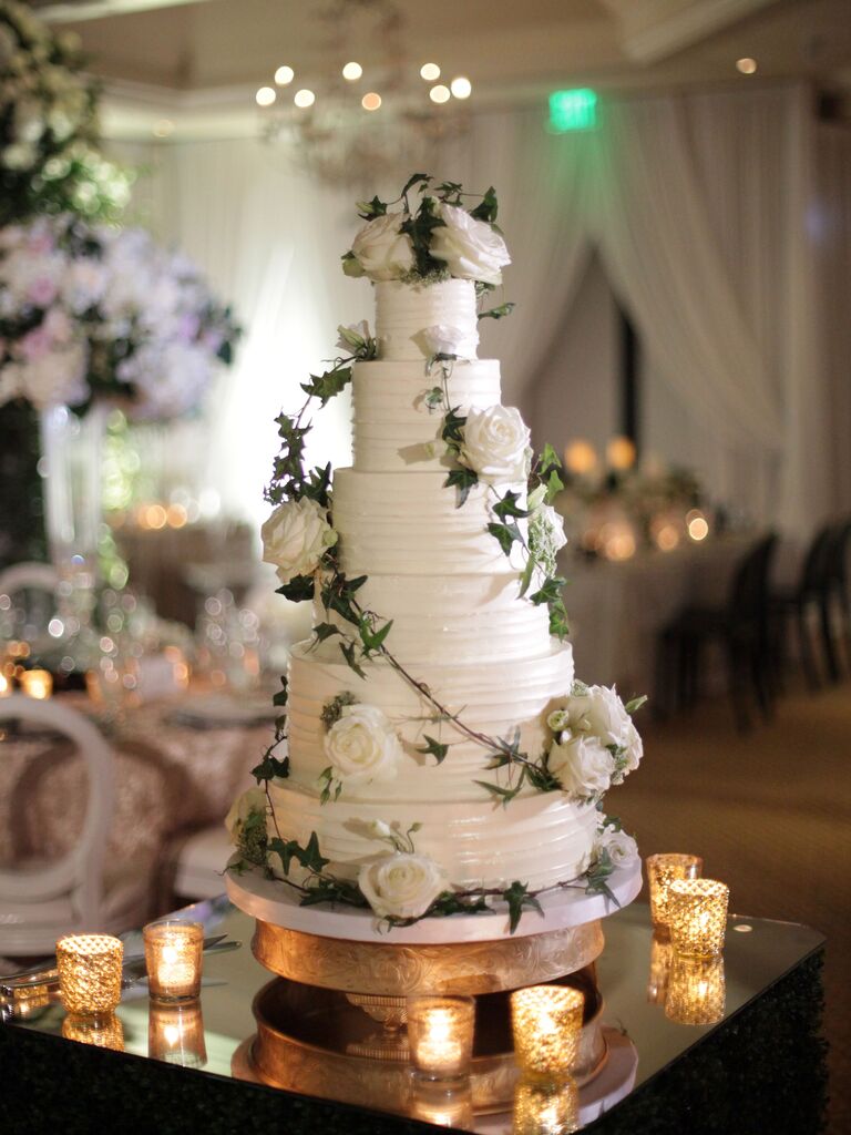 six tier wedding cake with white buttercream icing, ivy vines and white roses on a mirrored table surrounded by candles