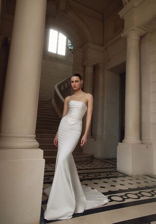Luminous satin defines the Lexi two-piece bridal set, offering an editorial approach to contemporary bridal style. The structured, strapless bodice features meticulous draping and ruching that wrap across the form, dipping into a subtle dropped waistline. Rendered in a choice of pristine ivory or light ivory, the design pairs this sculpted top with a fluid skirt that mirrors the bodice's artistic gathering. A striking slit opens along the front of the skirt, introducing dynamic movement and a flash of skin to counter the classic lines of the fabric. From the reverse, an open back concept balances the clean, architectural front. The skirt extends behind into a dramatic court train that trails naturally with every step, making it equally suited to a refined contemporary bridal look. Every angle of this Maison Blanche piece emphasizes texture and form for the bride seeking a fresh visual narrative.