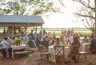 outdoor rehearsal dinner venue with guests seated at round tables with cross. back wooden chairs and string lights hanging above