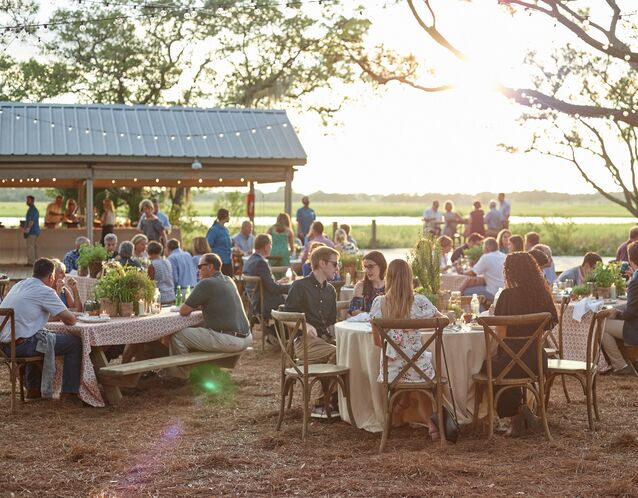 outdoor rehearsal dinner venue with guests seated at round tables with cross. back wooden chairs and string lights hanging above