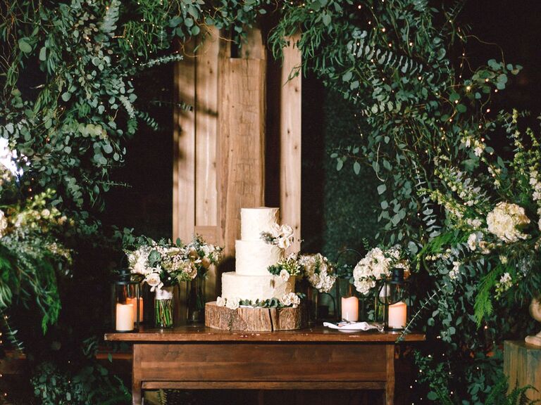 Three-tier rustic wedding cake on tree stump stand under greenery arch