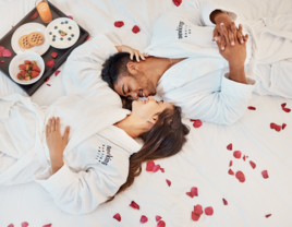 Couple laying on hotel bed in bath robes surrounded by rose petals and breakfast