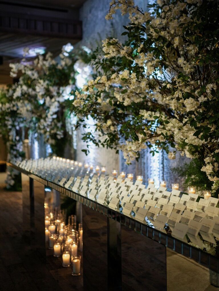 folded white wedding escort cards arranged on a mirrored tabletop with oversized white floral arrangements and votive candles
