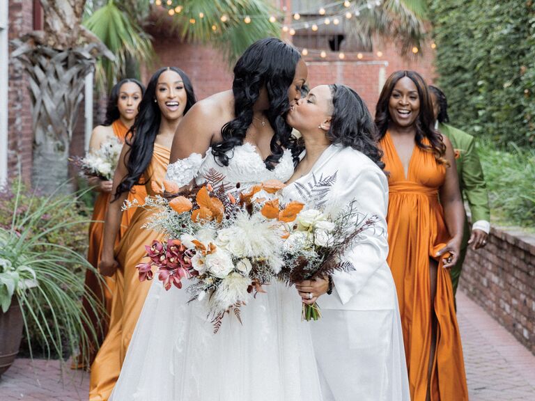 Two brides kiss while their bridesmaids cheer in the background