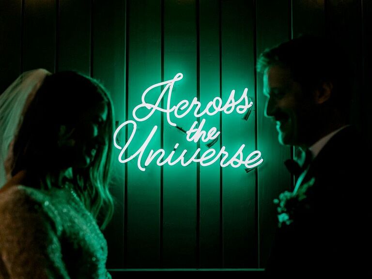 bride and groom standing out of focus in front of green neon sign that says across the universe