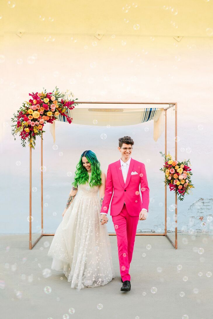 Bride with neon green hair and groom with hot pink suit walking away from copper chuppah.