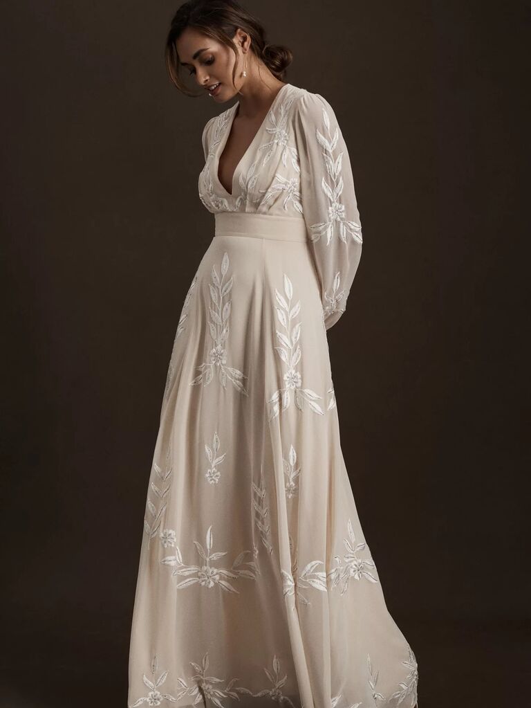 Model wears a bohemian cream-colored gown with white floral embroidery. 