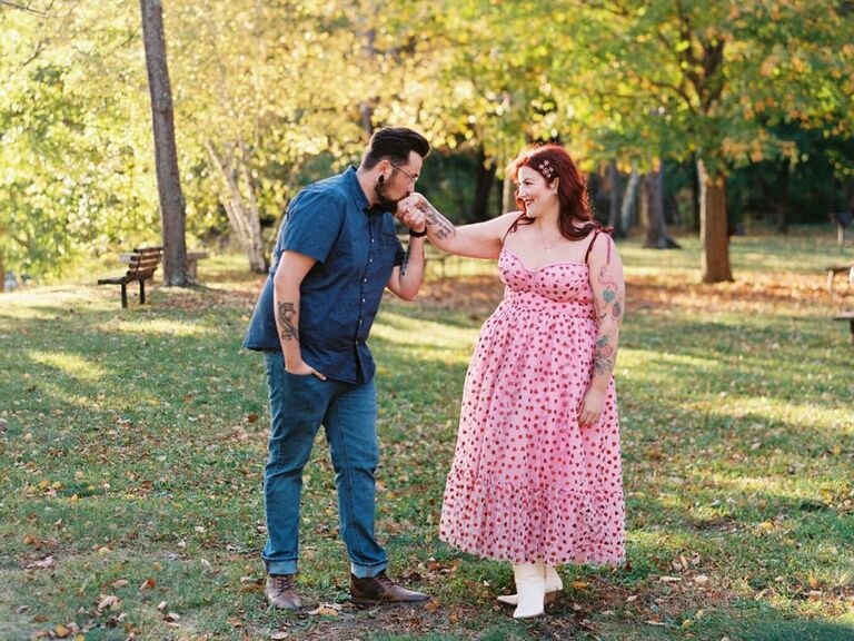 Man kisses his wife's hand while they stand in a beautiful leafy park. 