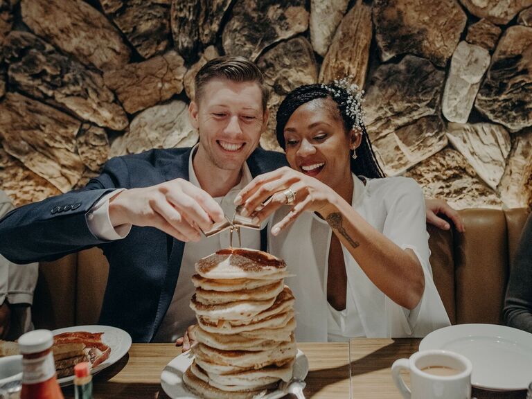 Couple pours syrup on a large stack of pancakes. 