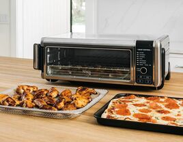 An airfryer sits beside plates of chicken wings and pizza.