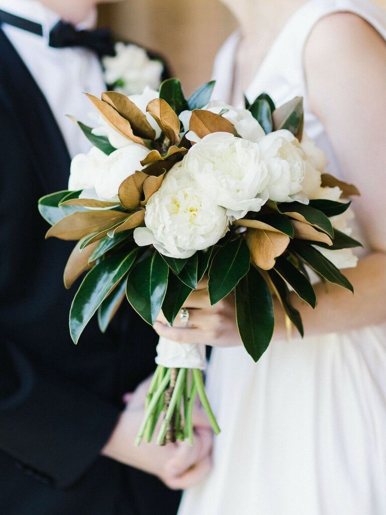 A couple hold a white peony bouquet with green and gold foliage.