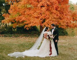 A couple pose in their wedding attire in front of a stunning autumn-toned tree.