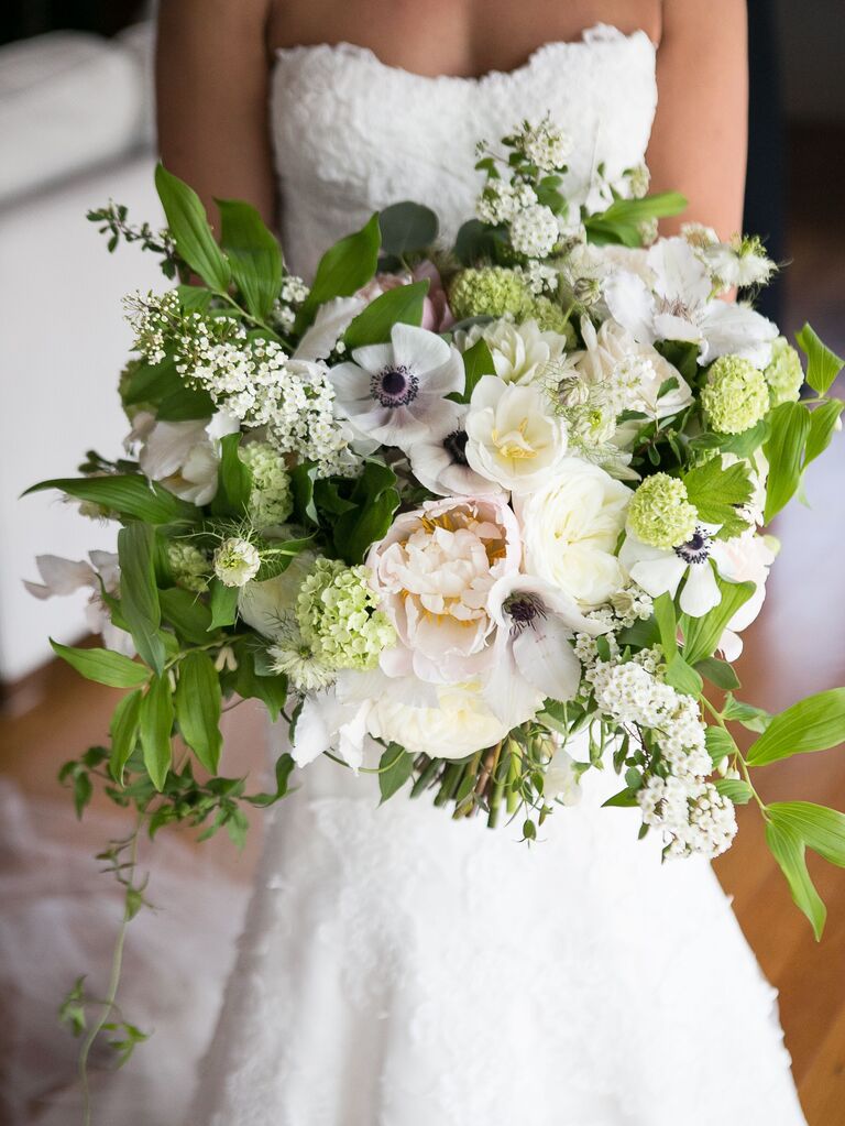 A bride holds an overflowing bouquet of peonies, anemones and greenery.