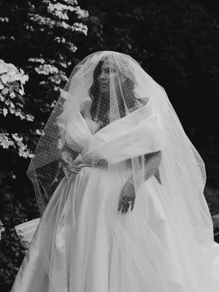 Model wears an elegant white wedding gown with a veil in front of her face. 