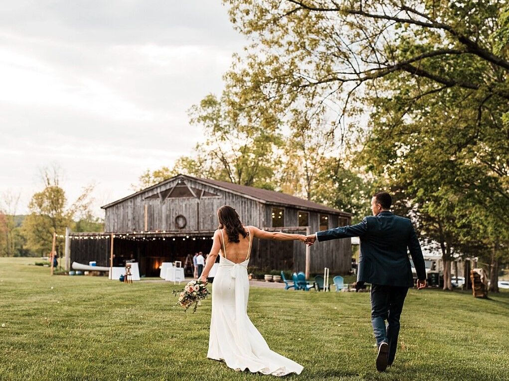 Couple holding hands walking towards the wooden barn venue with outdoor bistro lights
