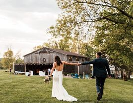 Couple holding hands walking towards the wooden barn venue with outdoor bistro lights