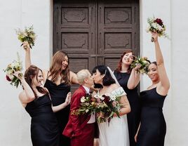 Bride with short hair and veil kissing spouse while surrounded by bridesmaids