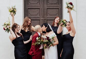 Bride with short hair and veil kissing spouse while surrounded by bridesmaids
