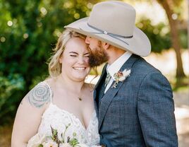 Man wearing a cowboy hat gives a sweet kiss on the cheek to his smiling bride. 