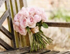 A pale pink bouquet sits propped, alone, against the back of a rustic wooden chair.