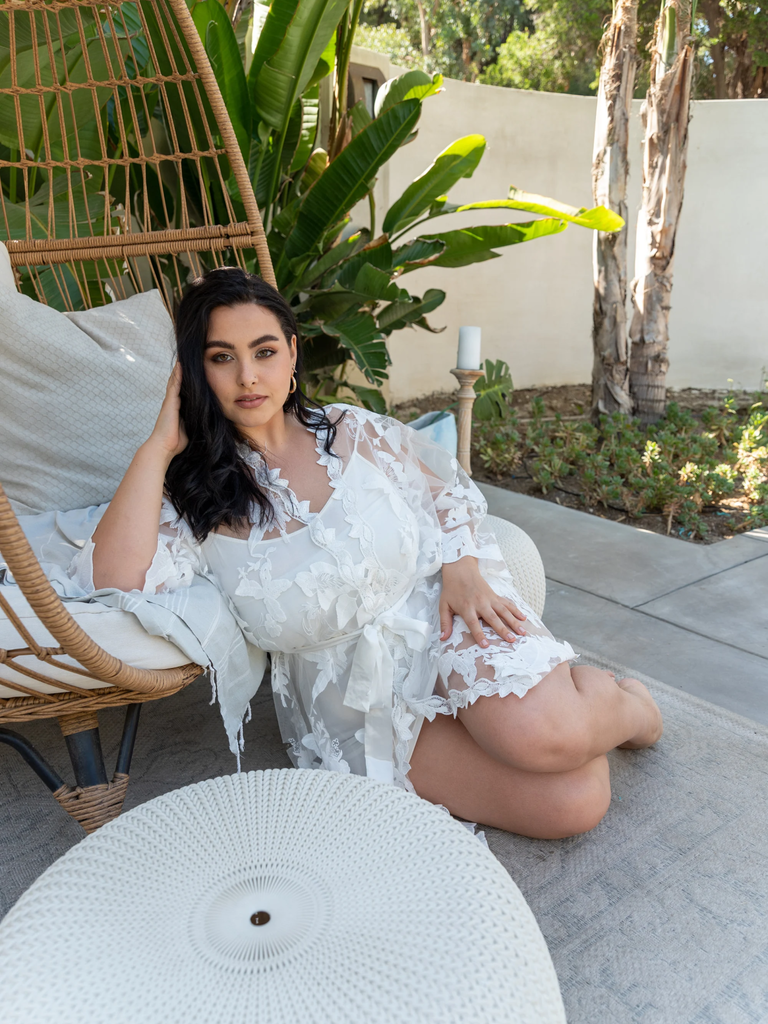 A model leans against a whicker chair wearing this Sheer Floral Bridal Robe.
