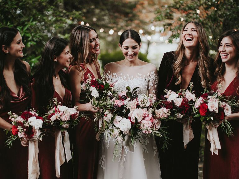 bride stands with bridesmaids wearing red velvet dresses and holding pink and dark burgundy bouquets with long ribbons