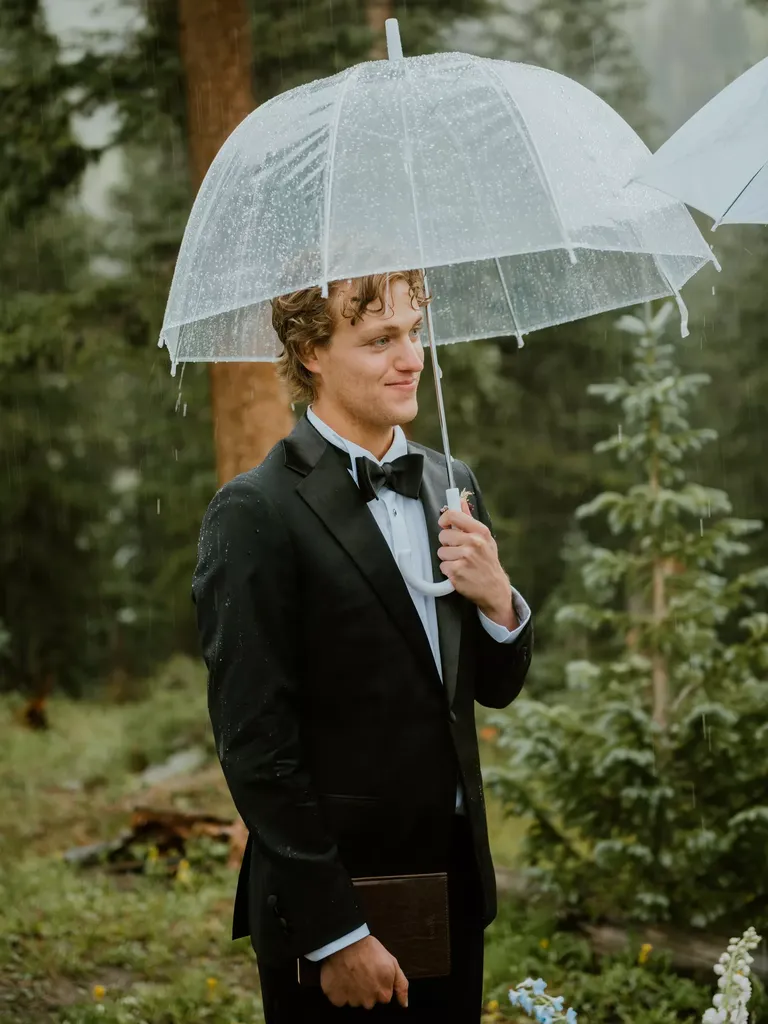 Groom in Classic Black Tuxedo Standing Under Clear Umbrella With Vow Book at Outdoor Ceremony
