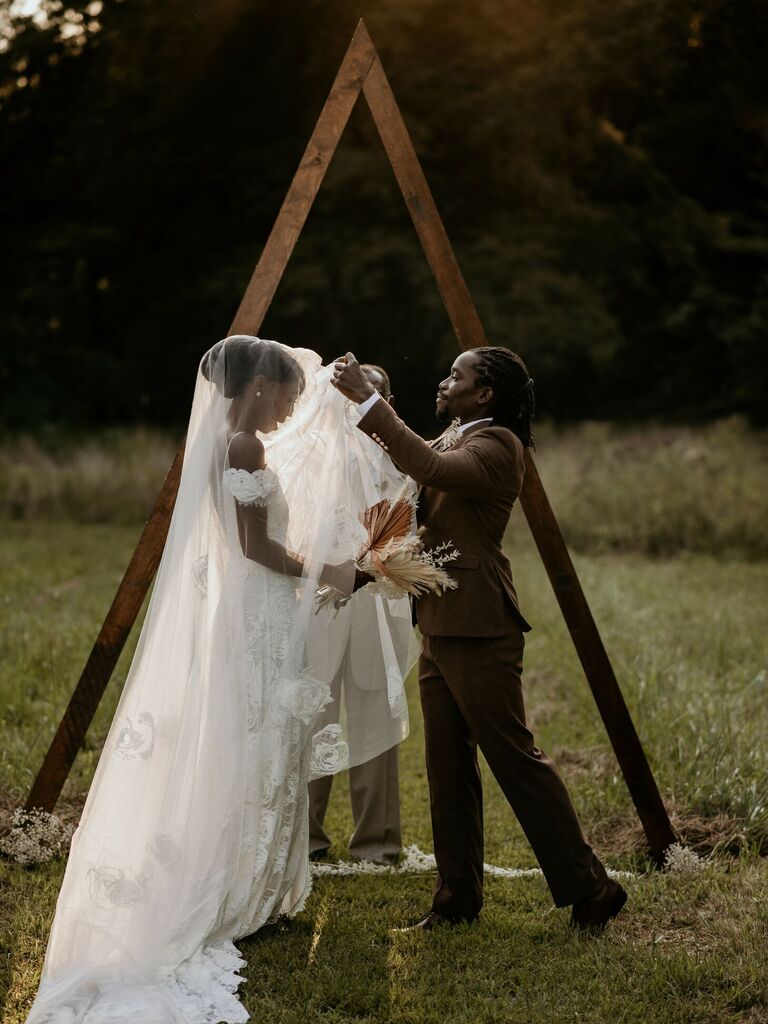Groom lifts bride's veil in front of rustic wood triangle arch