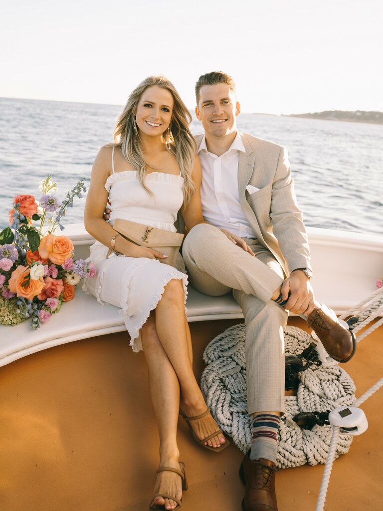 Couple sits on the bow of a sailboat while smiling at the camera. 
