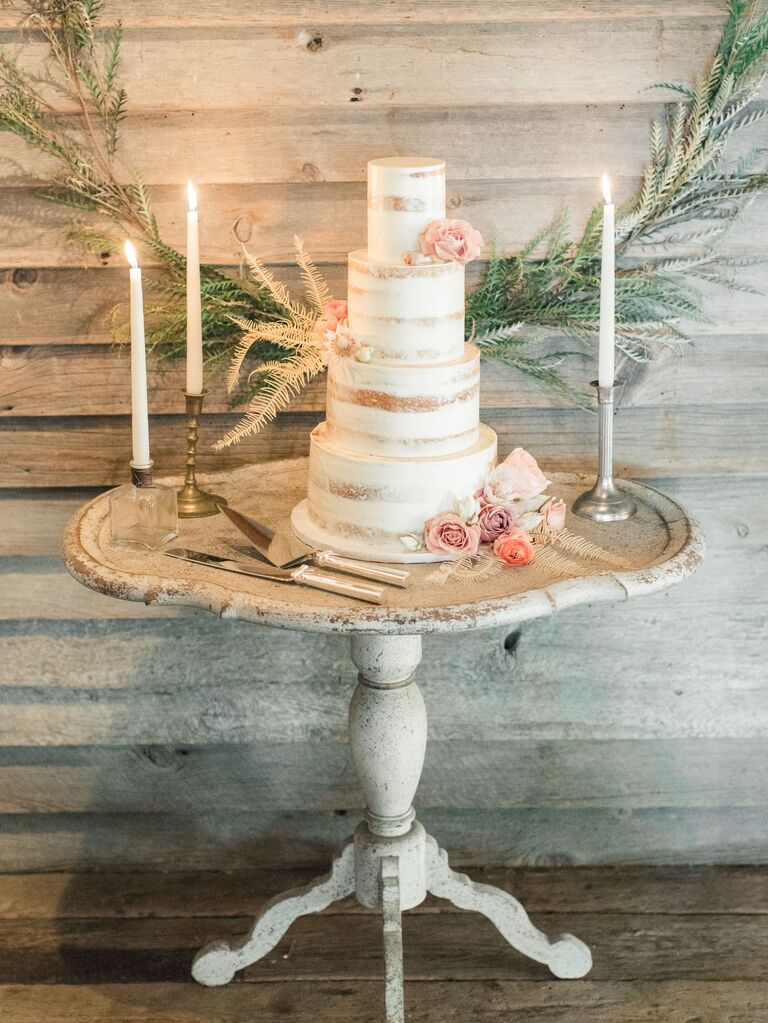 modern three tier semi naked wedding cake on whitewashed wooden table decorated with blush roses and bleached greenery
