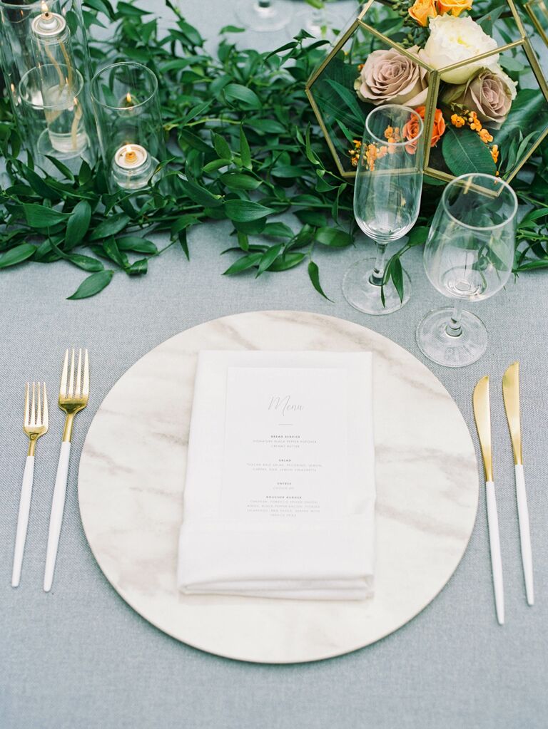 fall place setting with white marble charger, gray tablecloth, gold flatware and greenery table runner