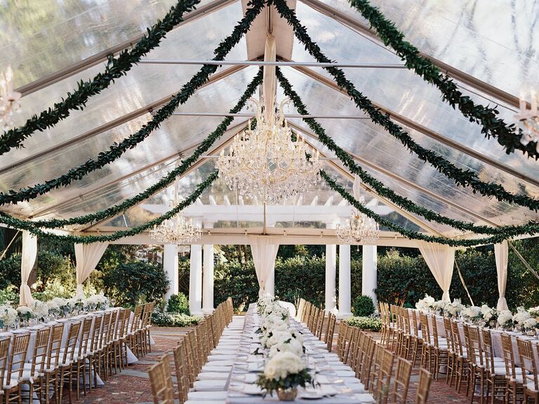 clear wedding reception tent with greenery garlands and chandeliers hanging from ceiling above long banquet tables