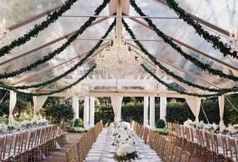 clear wedding reception tent with greenery garlands and chandeliers hanging from ceiling above long banquet tables