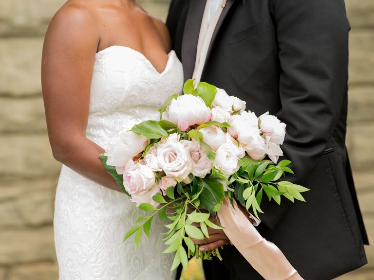 A couple stand together holding a white rose and peony bouquet.