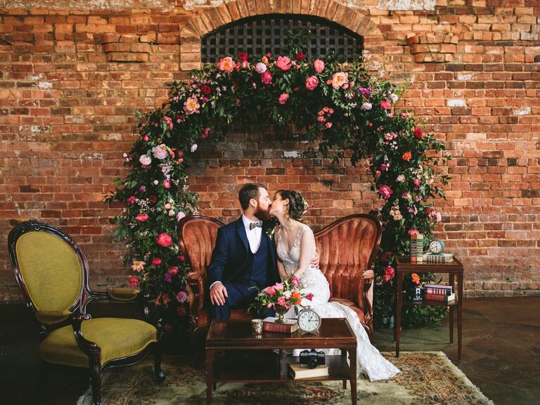 Couple sharing kiss while sitting on velvet couch in front of floral circle arch