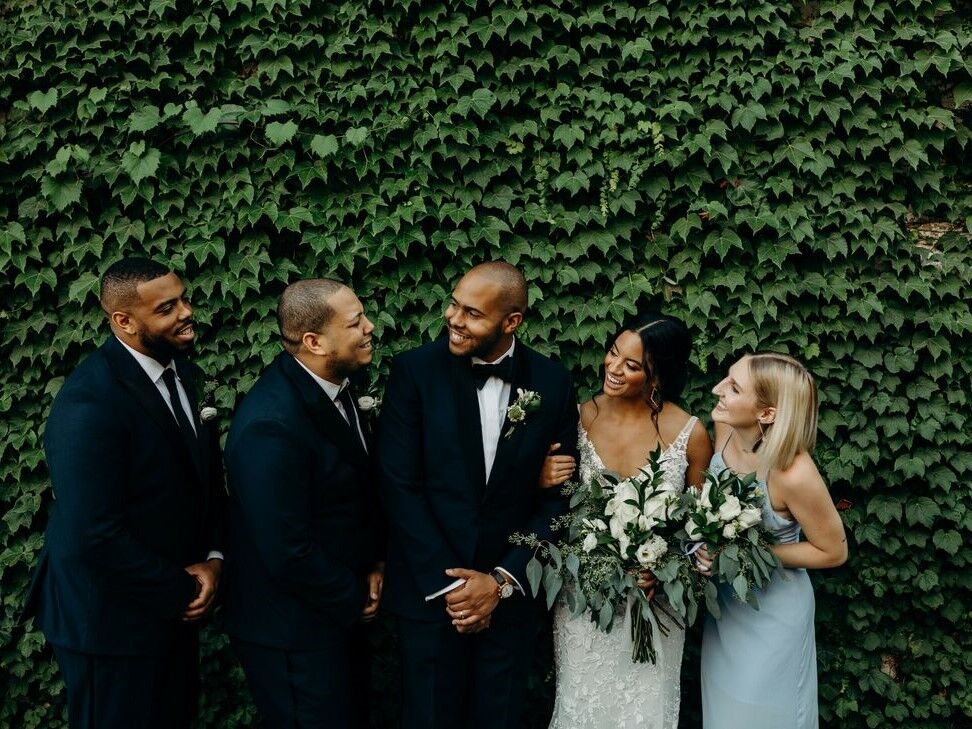 A groom poses with his bride and wedding party, leaning in for a laugh with his best man.