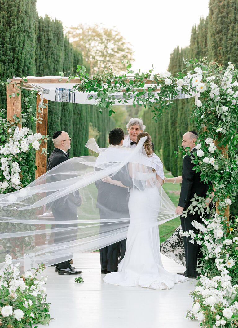 Couple under greenery-covered Chuppah with veil draped over them.