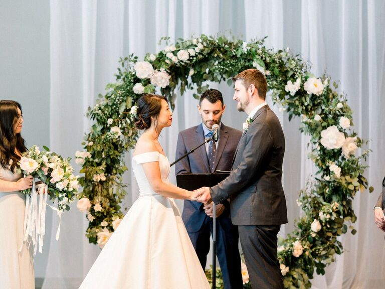 Couple holding hands and exchanging vows in front of white-and-green circle arch