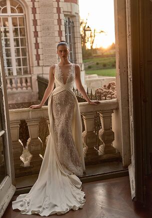 Satin and lace intertwine to create Reverie by WONÃ Concept. The fitted silhouette flares into a chapel or court train. Beadwork catches light across a structured bodice with a plunging illusion neckline, while a closed back maintains a classic impression. A detachable overskirt transforms the profile, accented by a matching bow. The ivory textile moves as the train follows in a luminous sweep.